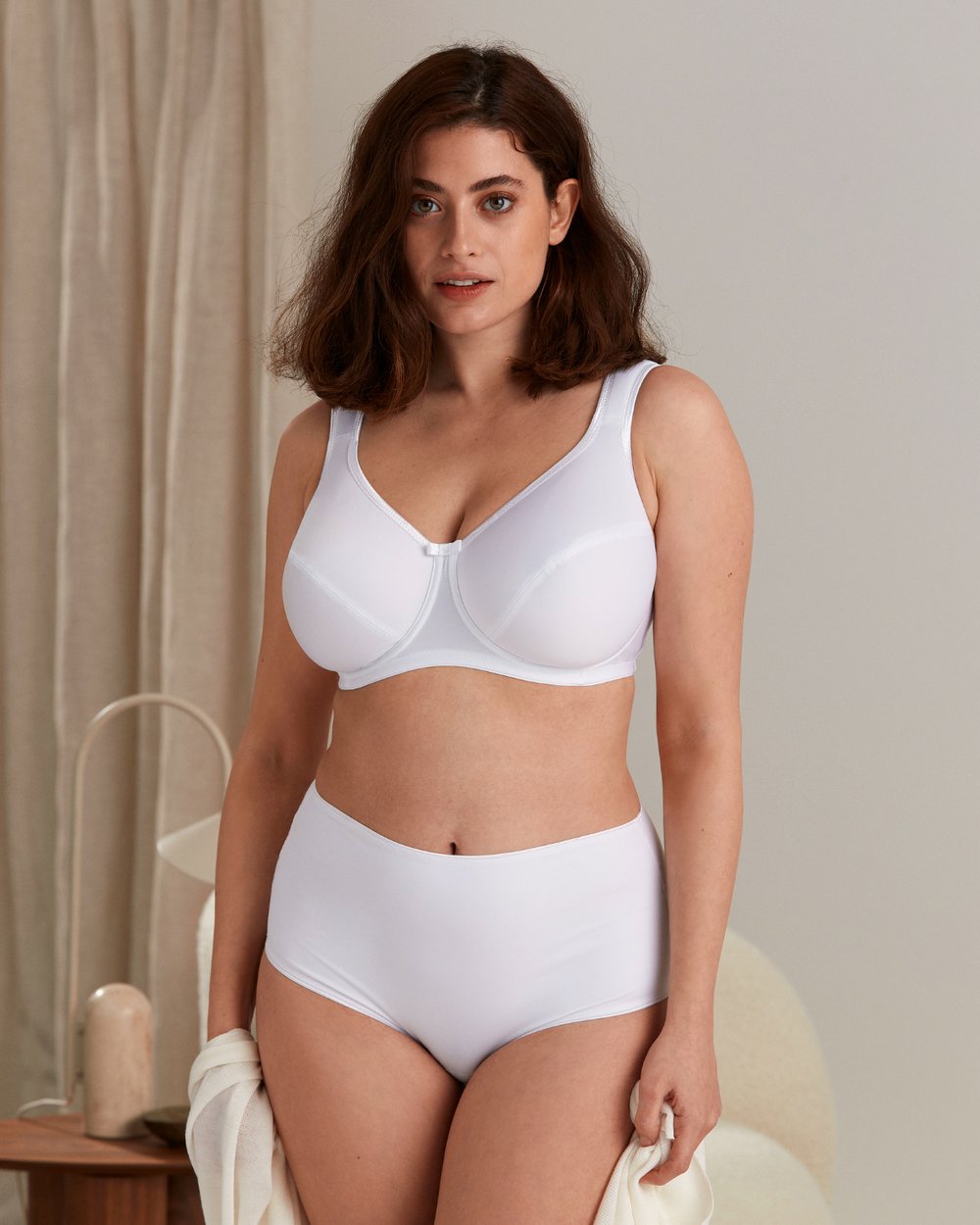 Person in a white bra and underwear stands indoors, holding a towel. A curtain and a small table are visible in the background.
