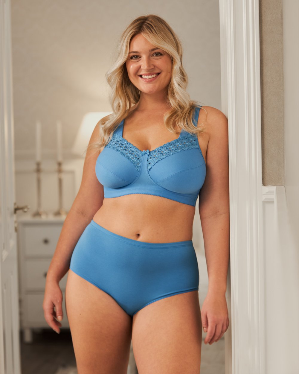 Smiling woman standing in a doorway wearing a blue lace-trimmed bra and high-waisted panties, with a well-lit interior background.