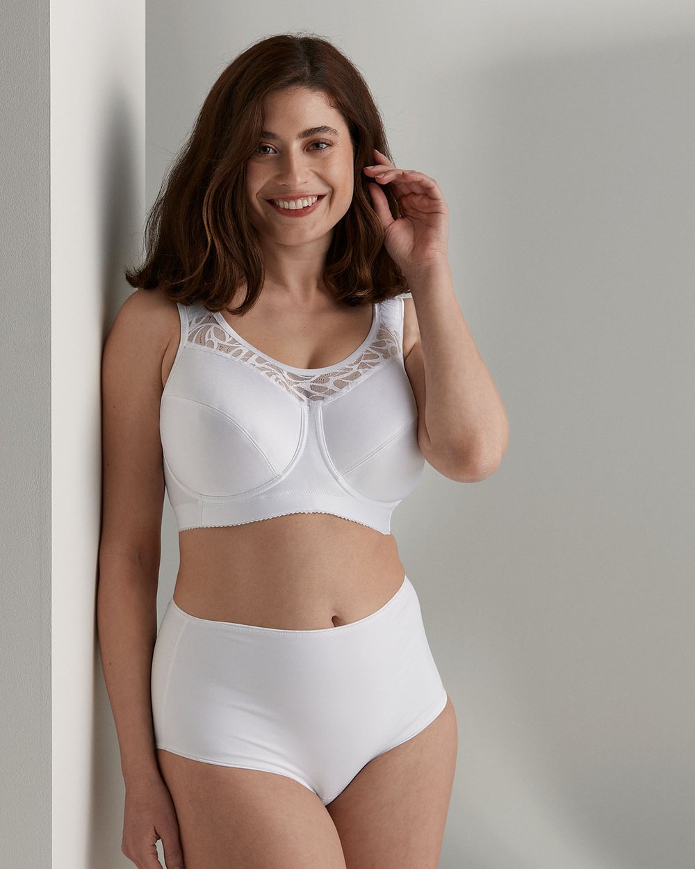 Woman in white lingerie smiling, standing in a bright room, touching her hair. She wears a lace-trimmed bra and high-waisted briefs.