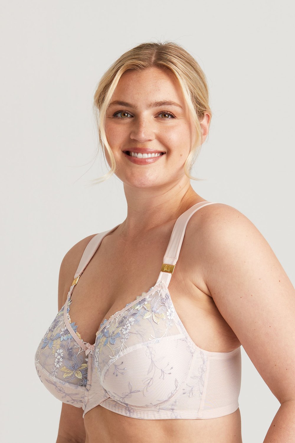 Smiling woman wearing a pale pink floral embroidered bra, blonde hair pulled back, on a neutral background.
