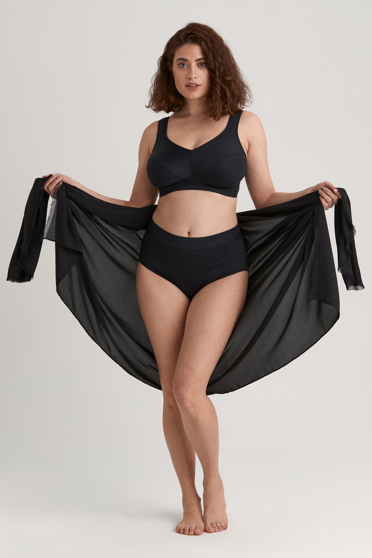 Femme tenant ouvert un sarong léger noir Miss Mary aux bords frangés, porté avec un maillot de bain noir.