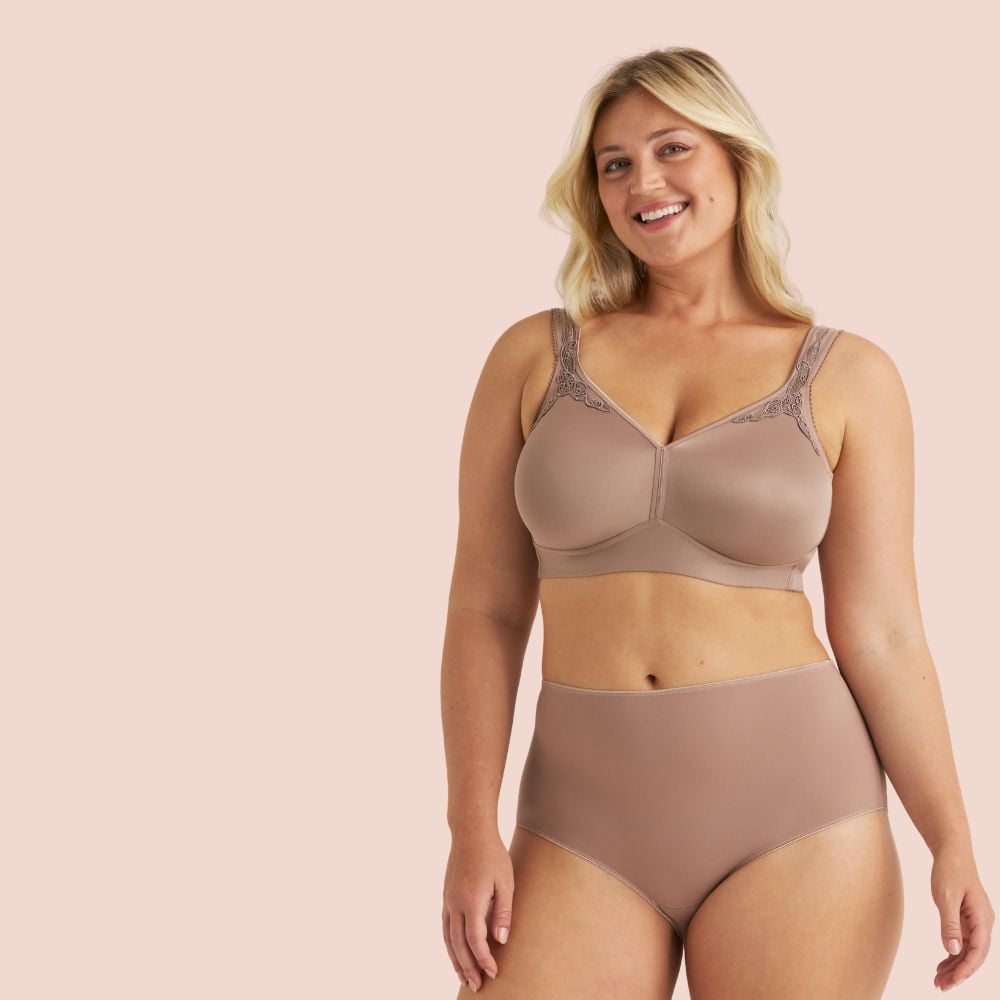 A smiling woman wearing a matching beige bra and high-waisted underwear, standing against a light pink background.