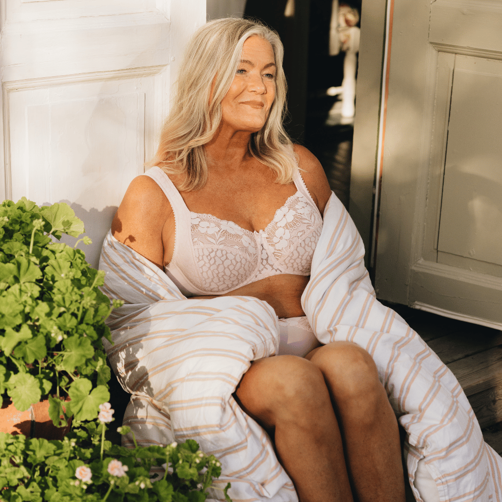 A smiling woman sits wrapped in a striped blanket, wearing lace lingerie, with sunlight casting soft shadows, next to potted plants by an open door.