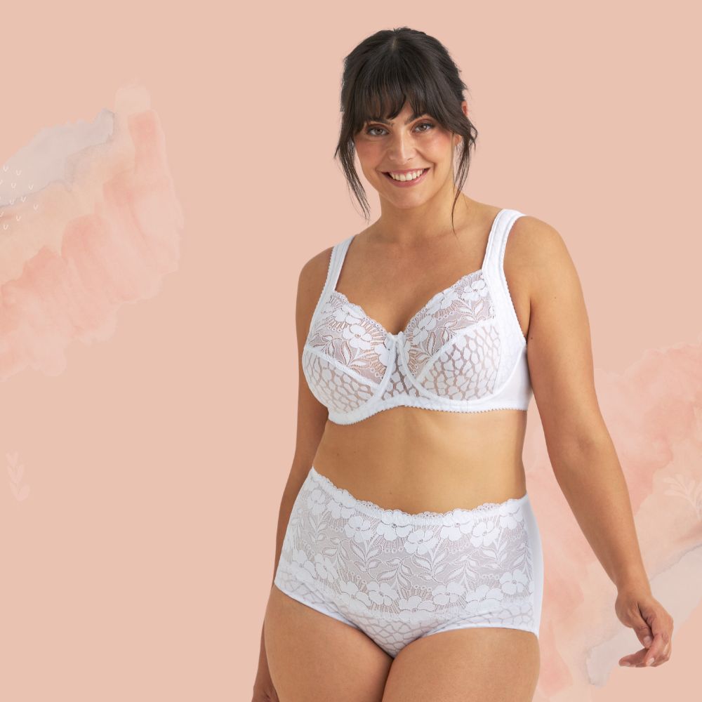 A woman smiling, wearing a white lace bra and matching high-waisted panties, standing against a soft pink background.