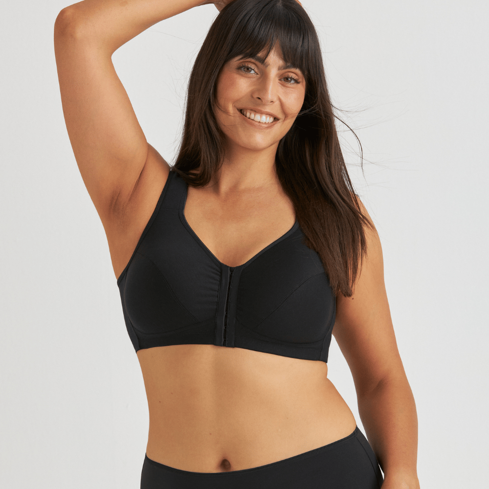 A woman with long dark hair wearing a black bra smiles with her left arm raised against a plain background.