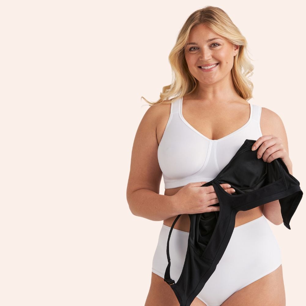 A woman smiling, wearing a white bra and underwear, holding a black garment against a light background.