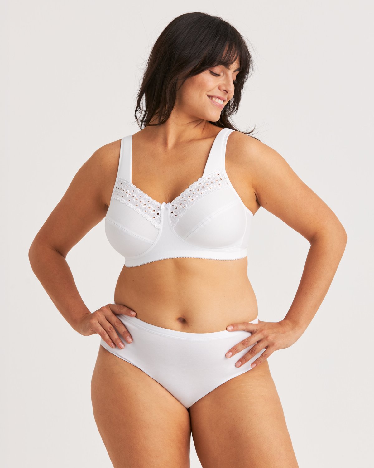 Woman in a white bra with lace detailing and white underwear, smiling and looking to her left against a plain background.