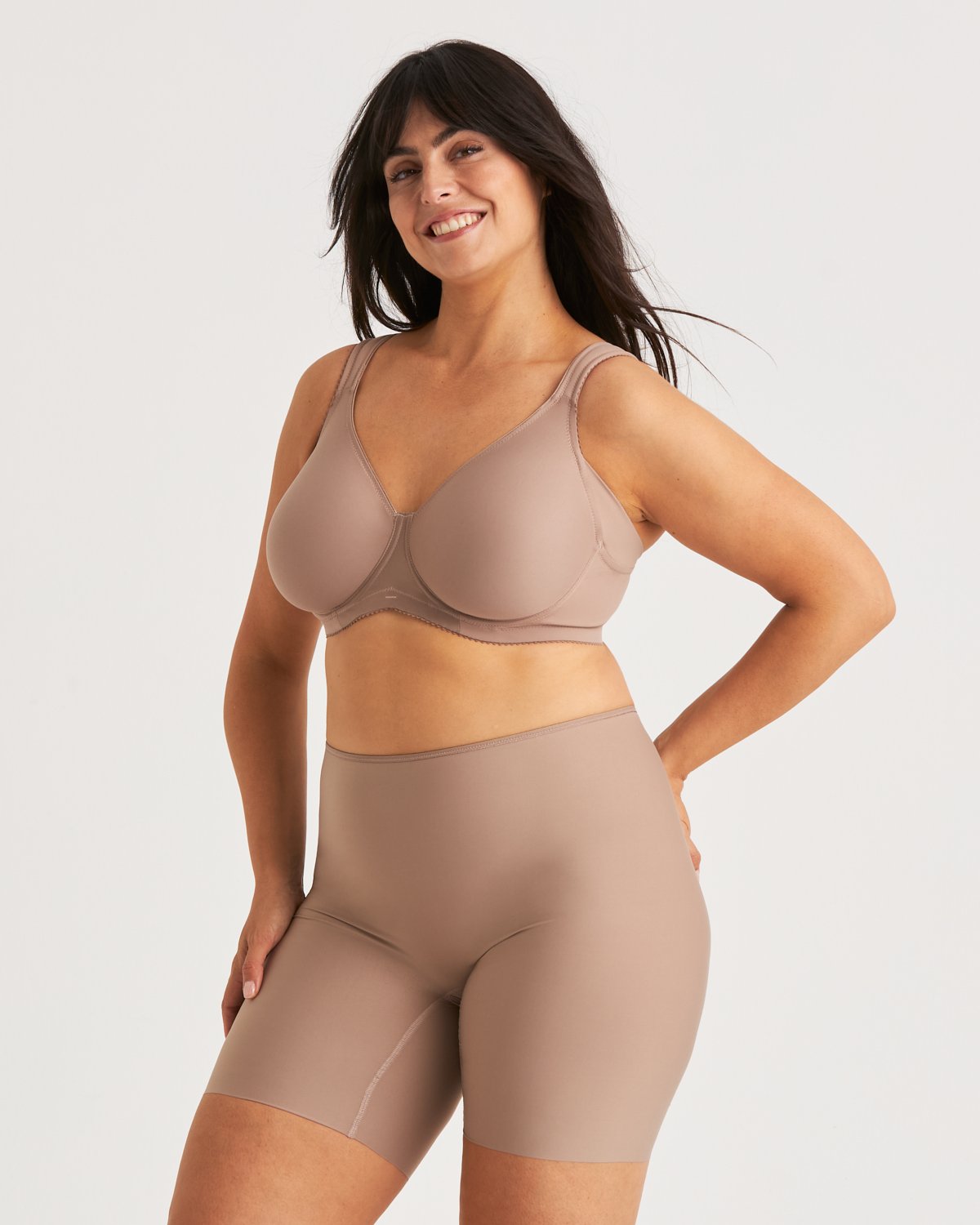 Smiling woman in a taupe bra and high-waisted shorts, posing confidently against a plain background.