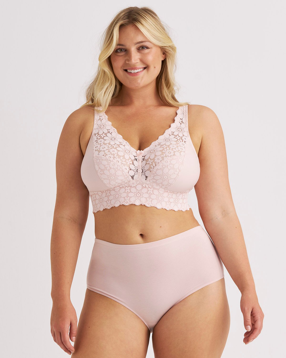 Smiling woman in pink lace bralette and high-waisted underwear against a plain background.
