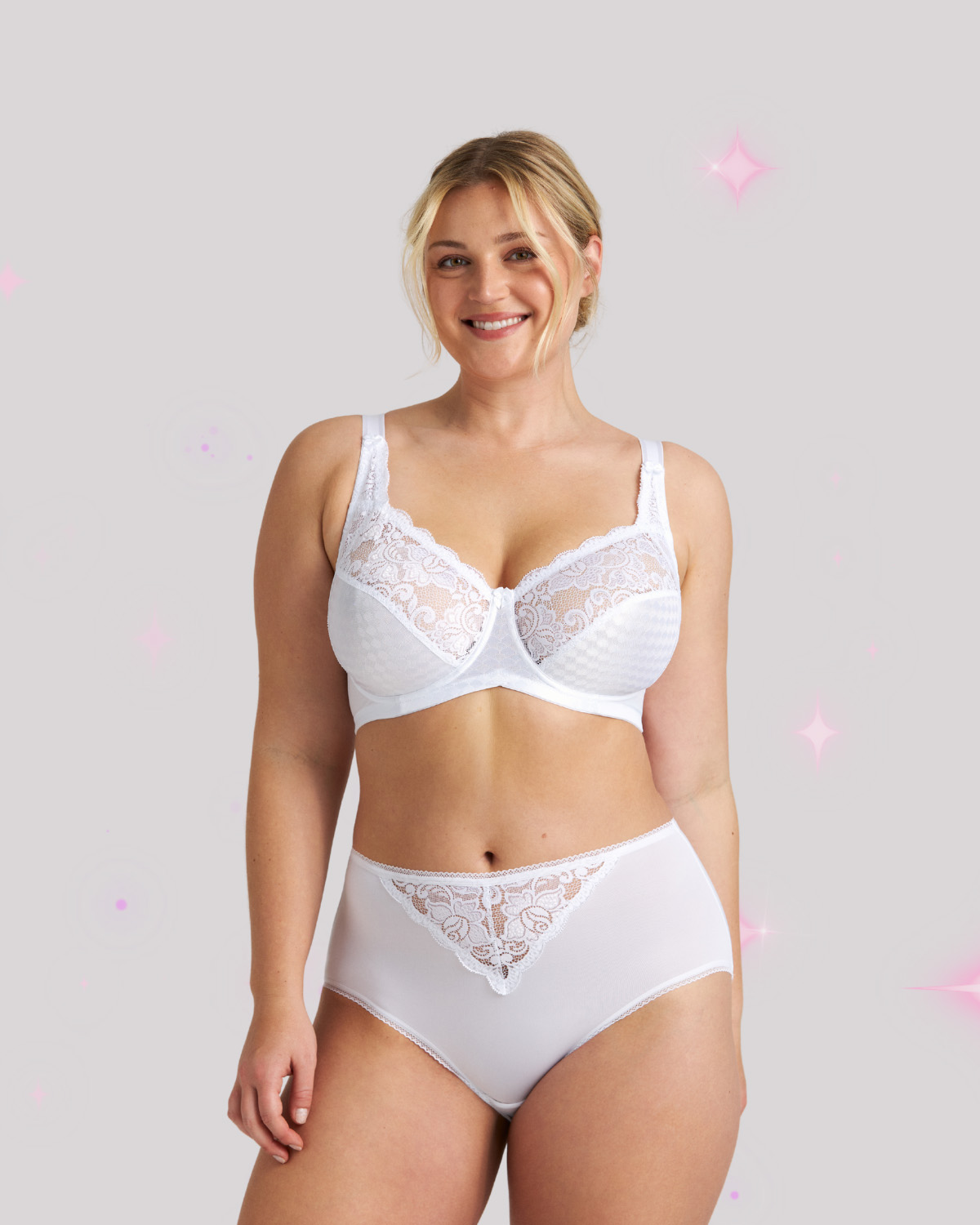 Smiling woman modeling white lace bra and matching high-waist panties against a pale background with subtle pink sparkles.