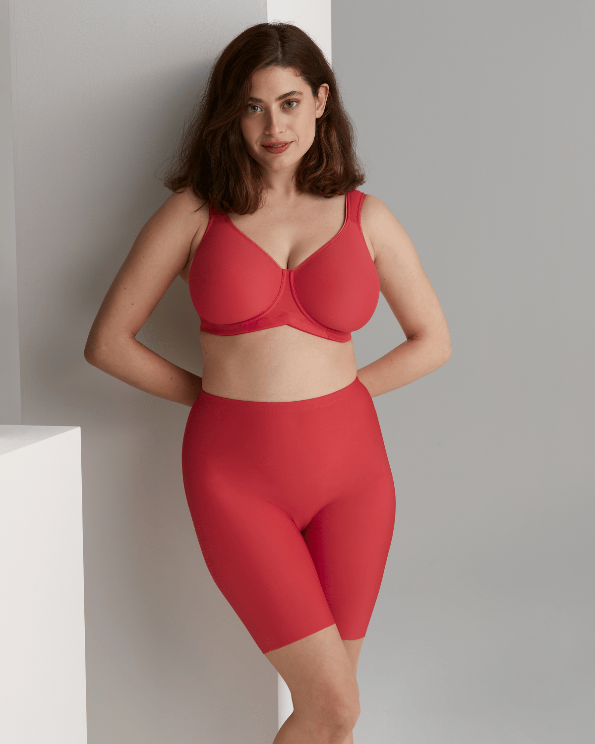 Woman in red shapewear stands against a wall, with arms behind her back, looking confidently at the camera in a minimalist setting.
