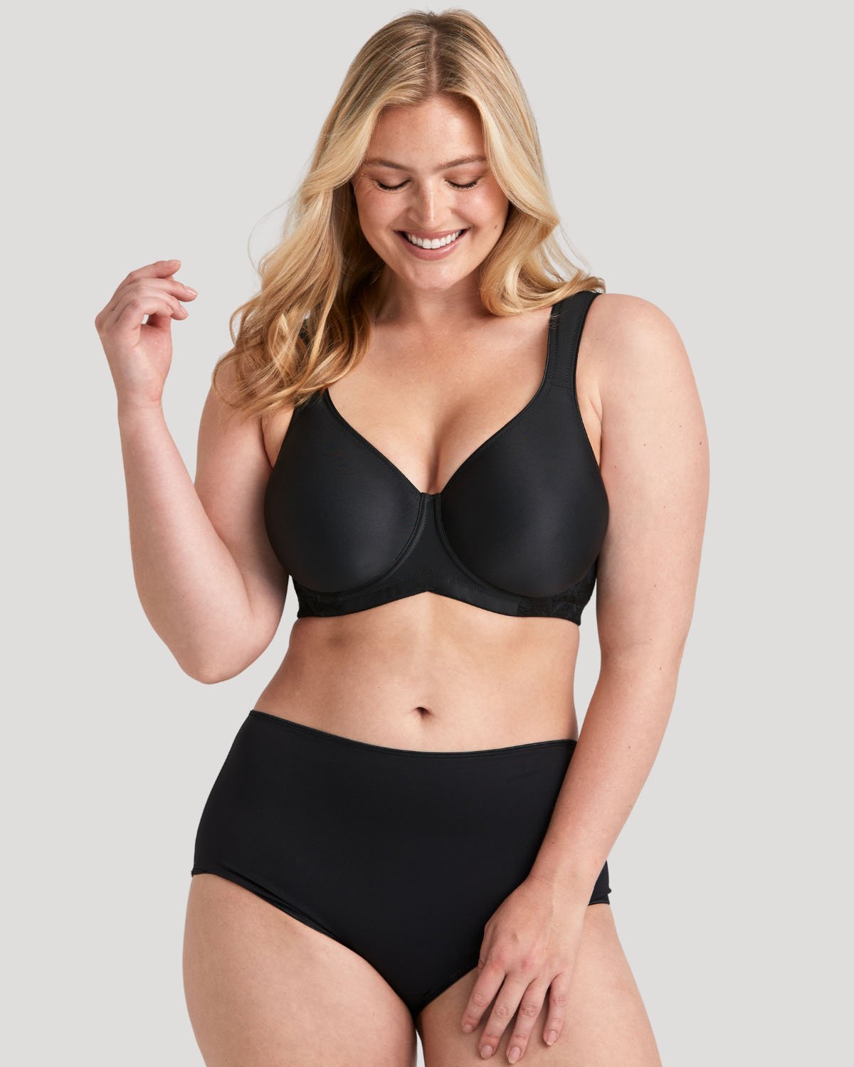 Smiling blonde model in black underwire bra and high-waisted briefs, posing against a light gray background.