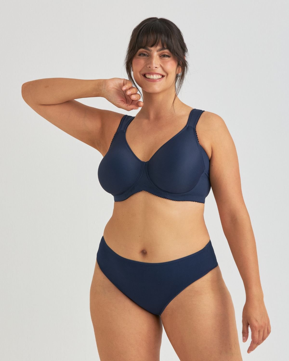A smiling woman poses confidently in a navy blue bra and panties against a plain background.