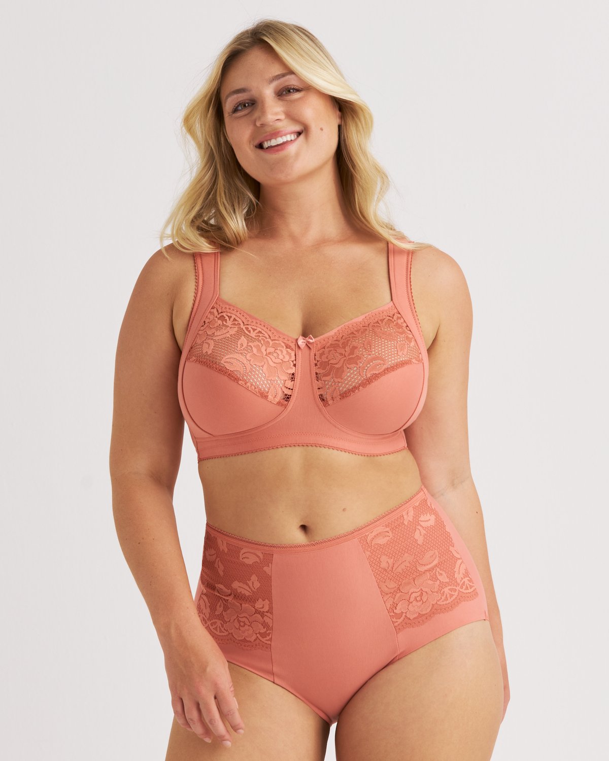 A woman smiles while wearing a matching peach-colored bra and high-waisted panties with lace details.