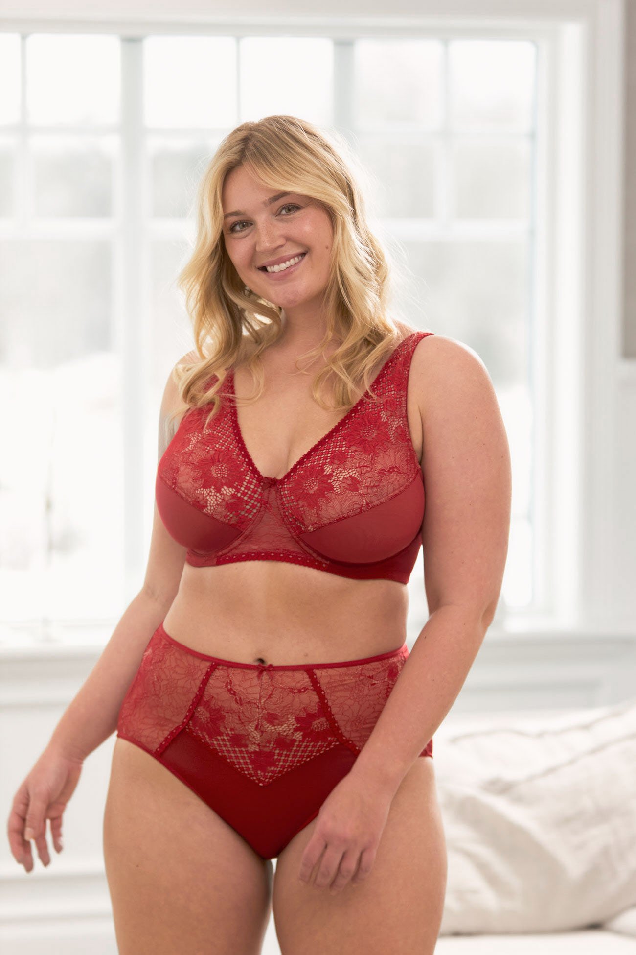 Woman smiling in a bright room, wearing red lace lingerie with floral patterns, standing in front of a window.
