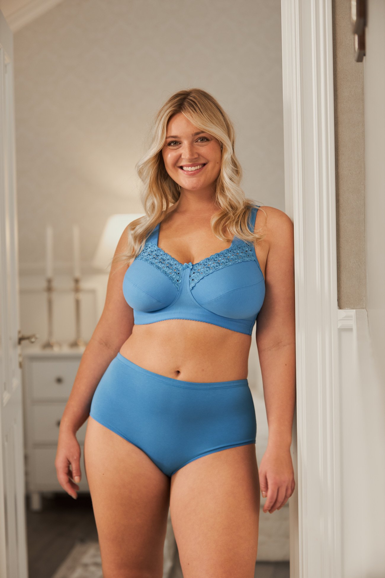 A woman with blonde hair wearing a blue lace-trimmed bra and briefs, smiling in a softly lit room.
