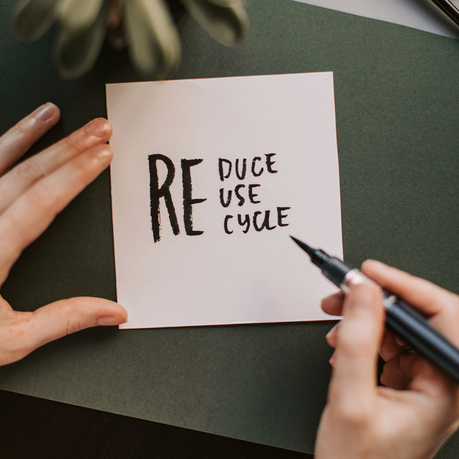Hands holding a card with handwritten text: "Reduce, Reuse, Recycle" on a green surface, with a pen and a plant nearby.