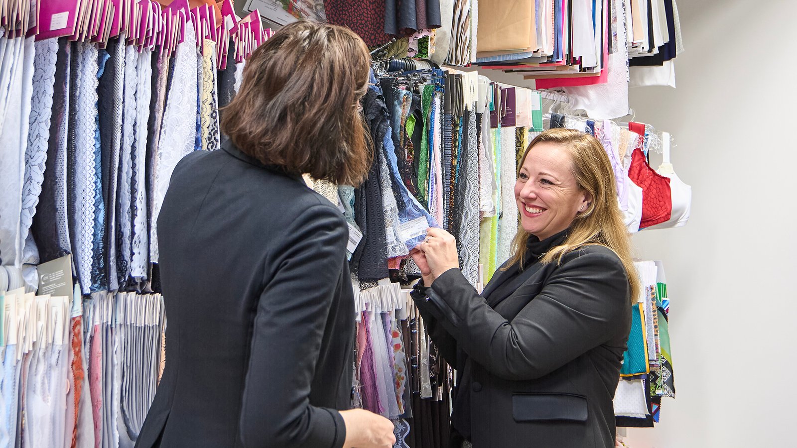 Two women in a fabric store, one with brown hair pointing at fabric rolls, while the other, blonde, smiles and holds a fabric sample.