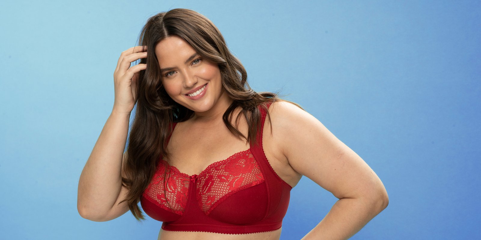 Woman in a red lace bra smiles, touching her hair, against a blue background.