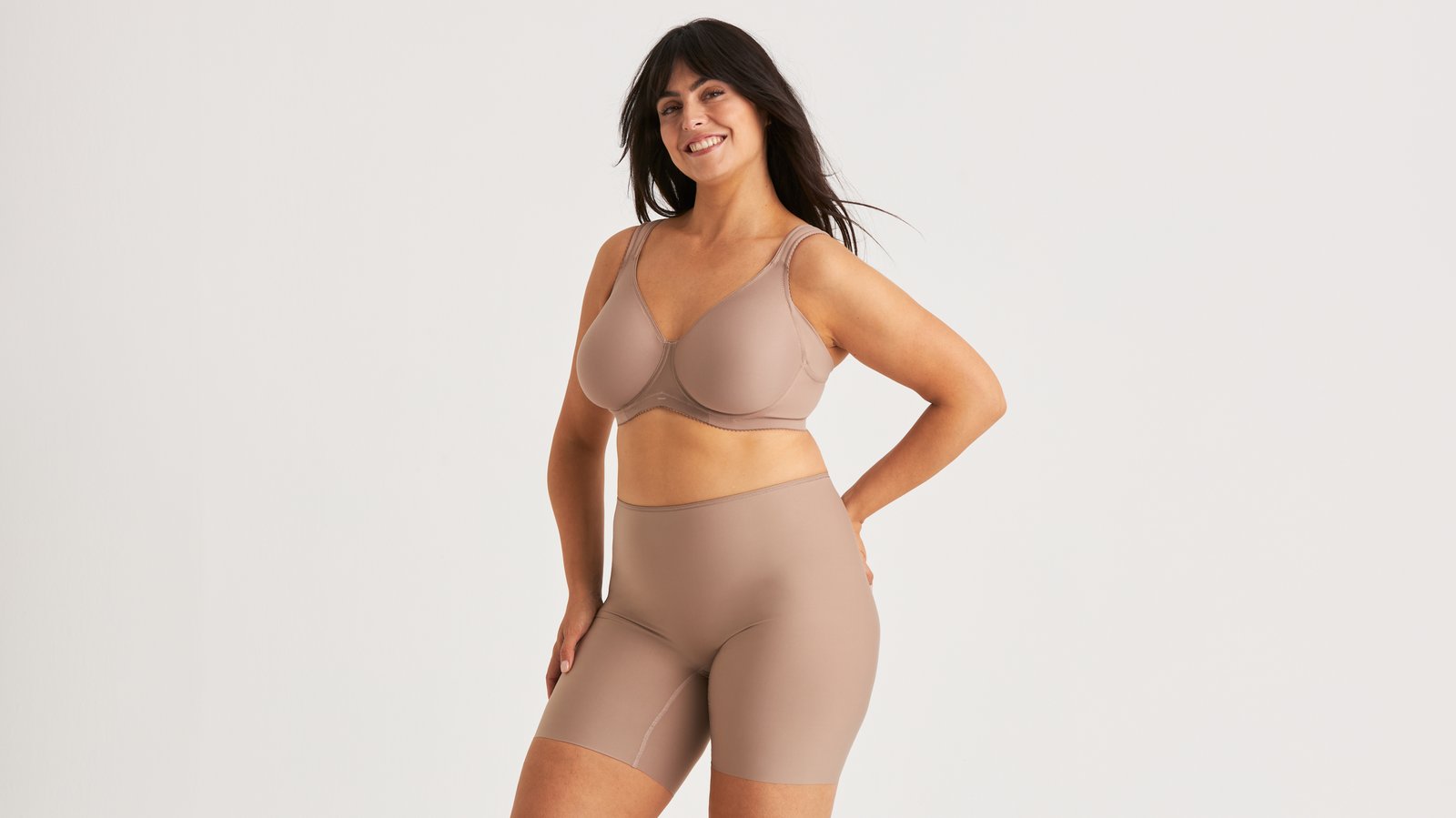 A woman smiling, wearing a taupe-colored bra and high-waisted shorts, standing against a plain white background.