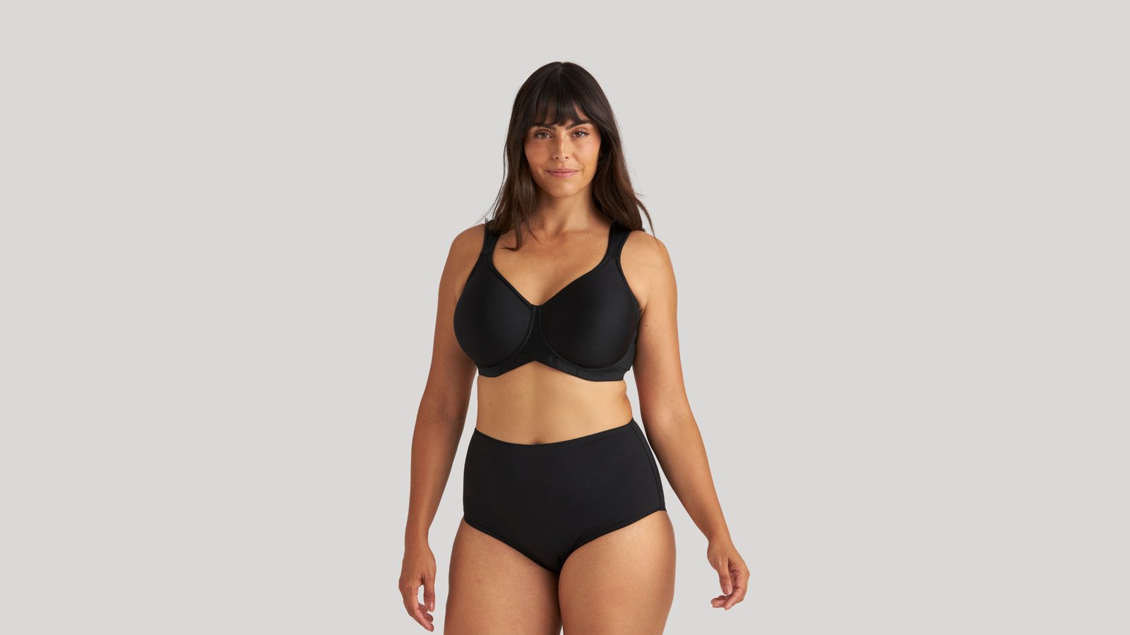 Woman wearing a black bra and high-waisted panties, standing confidently against a plain gray background.