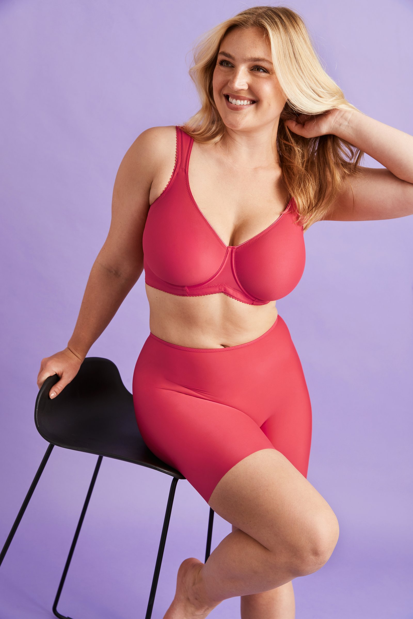 A smiling woman in a pink bra and shorts sits on a black chair against a light purple background, touching her hair.