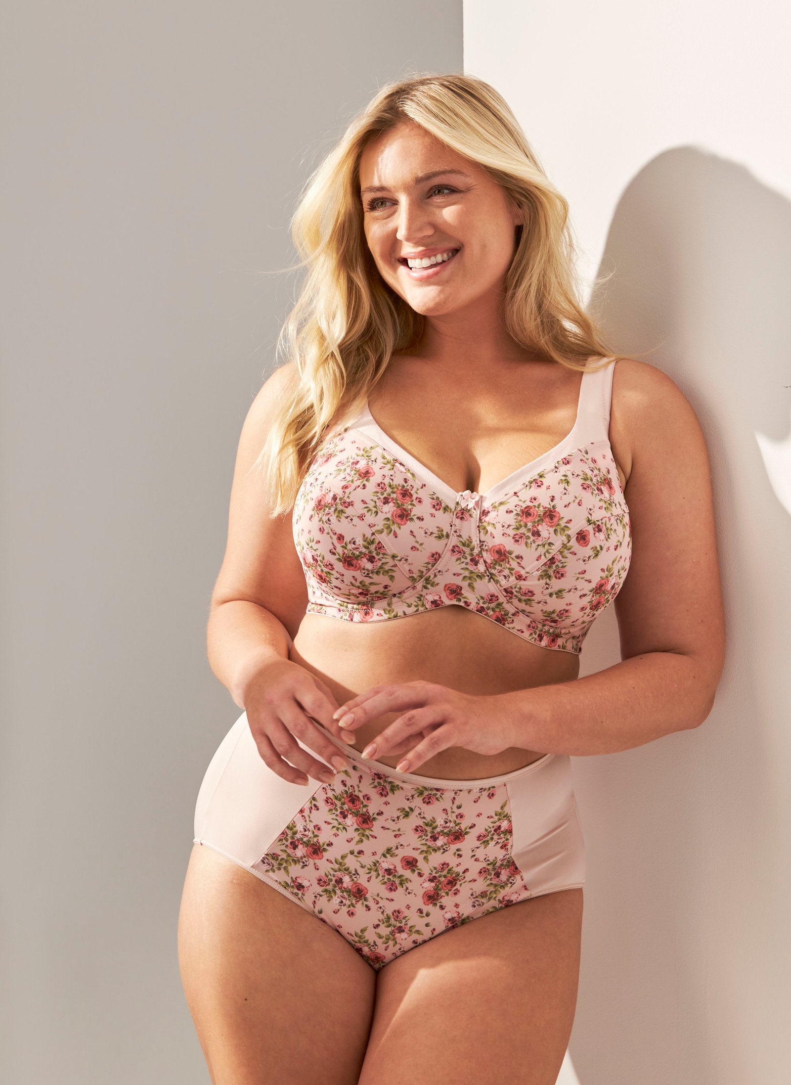 Woman wearing a floral pink lingerie set, smiling and standing against a white wall, bathed in soft natural light.