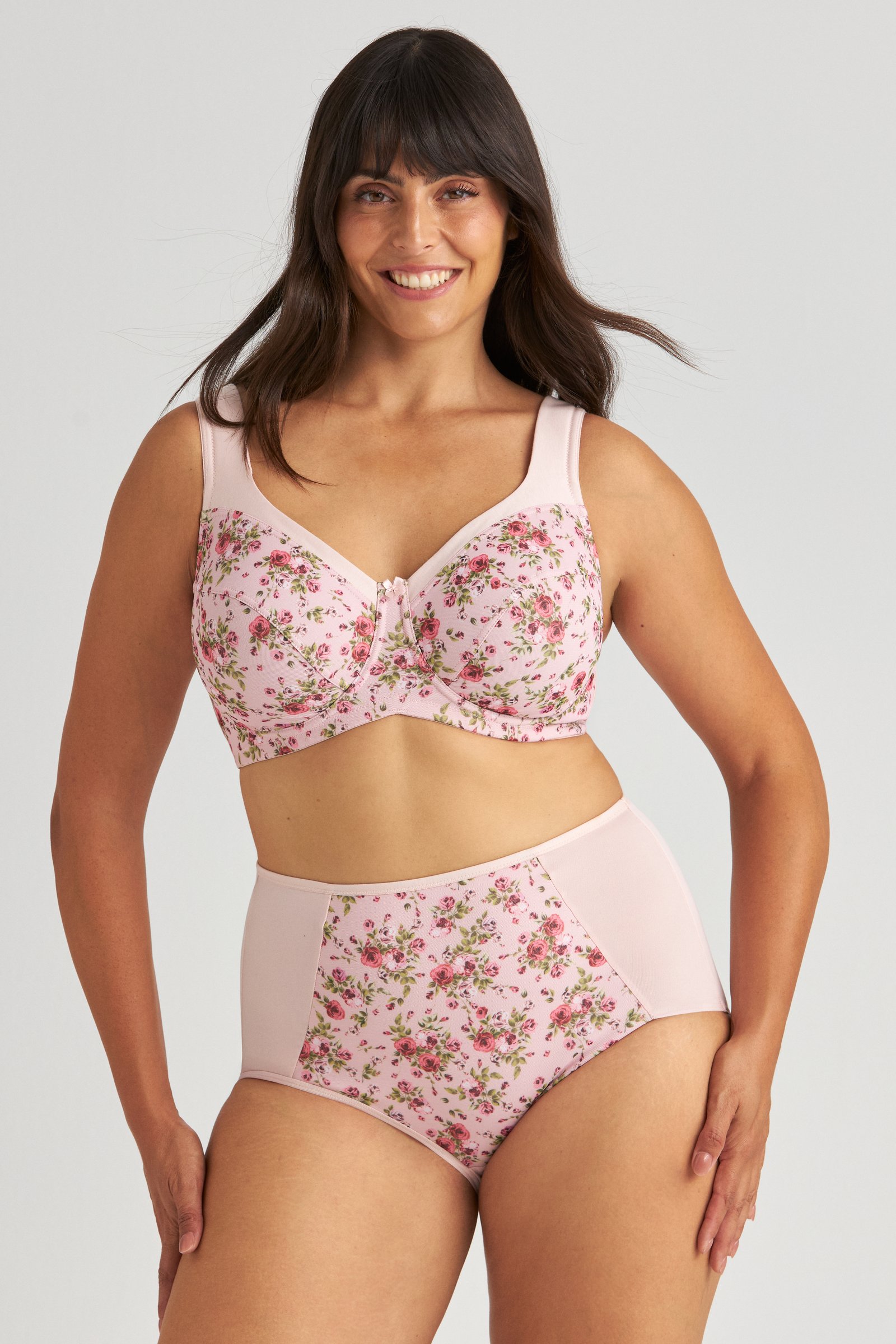 Smiling woman in floral pink lingerie with a supportive bra and high-waisted briefs, standing against a light gray background.