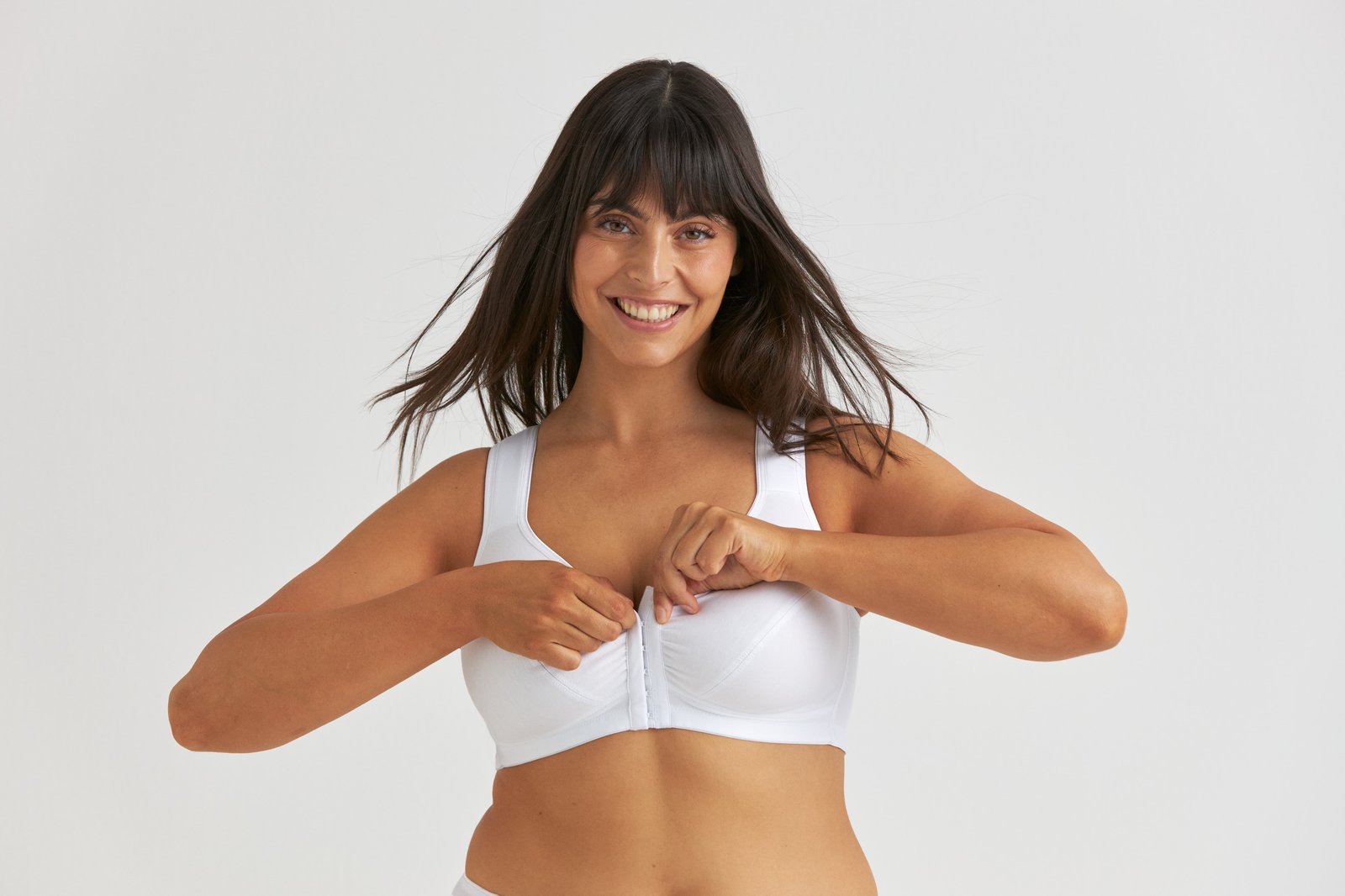 A woman smiling, fastening a front-closure white bra, with dark hair and a neutral background.