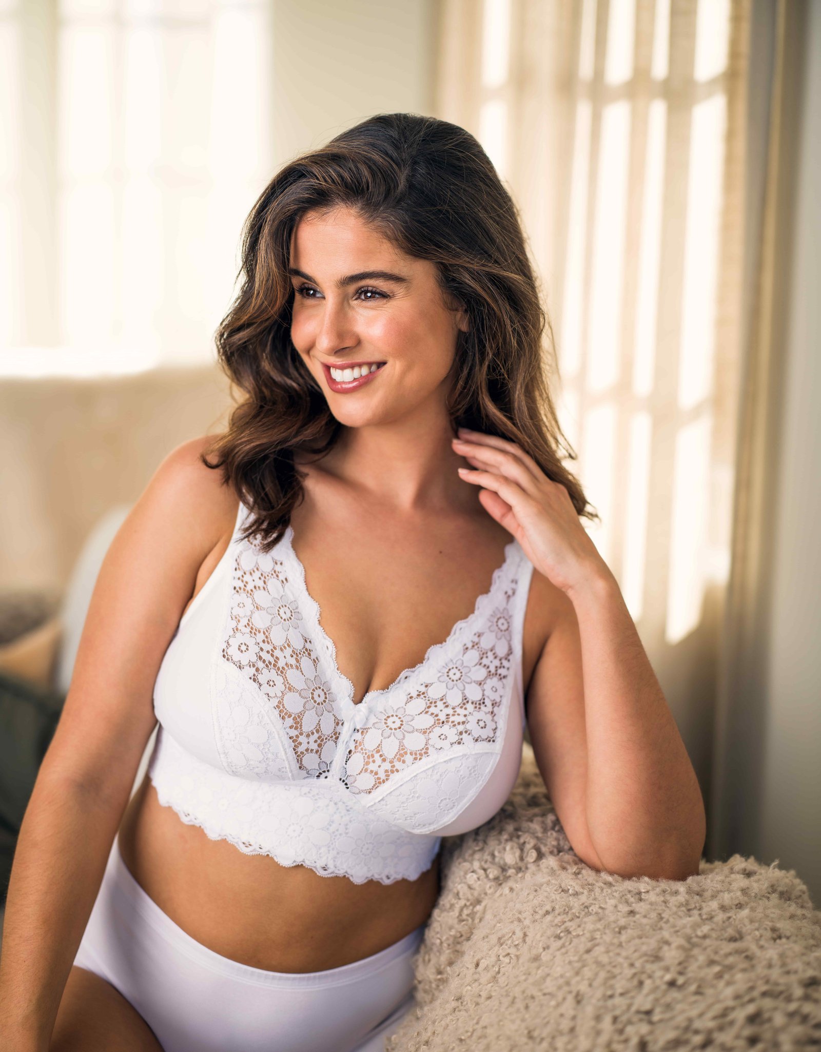 A woman in a lace-trimmed white bralette and underwear smiles while seated on a cozy beige couch, with a softly lit background.